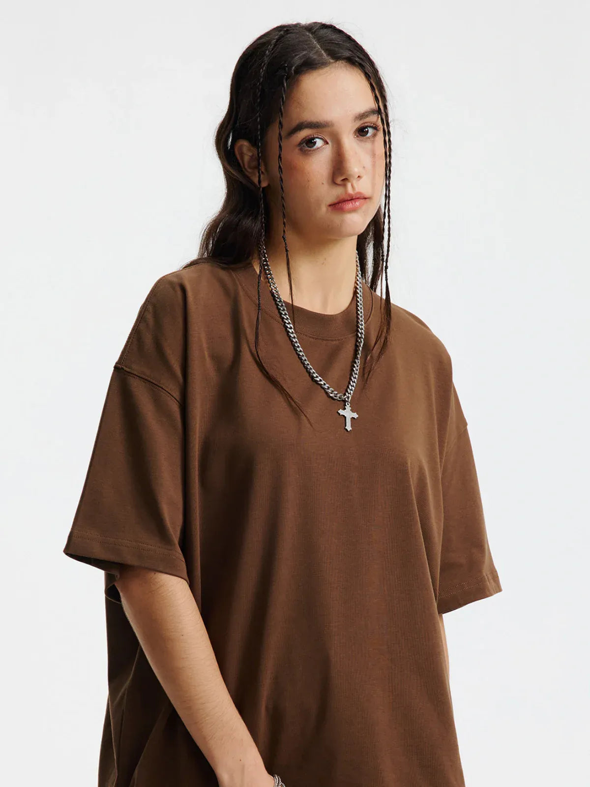 Person with long dark hair in braids wears the Custom Anime Character Chocolate Brown T-Shirt—Personalized 230GSM 100% Cotton Tee—and a silver chain cross necklace, standing against a plain white background.