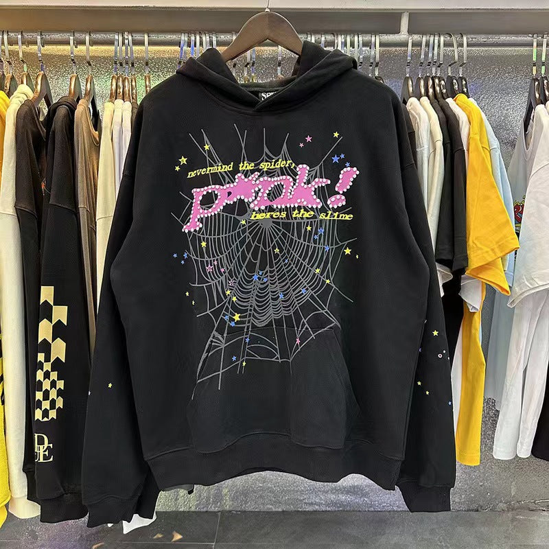 A Sp5der Trendy Hoodie in black with a vibrant web print and &quot;Prink!&quot; in bold pink letters is displayed on a rack, exuding streetwear vibes, surrounded by colorful t-shirts.