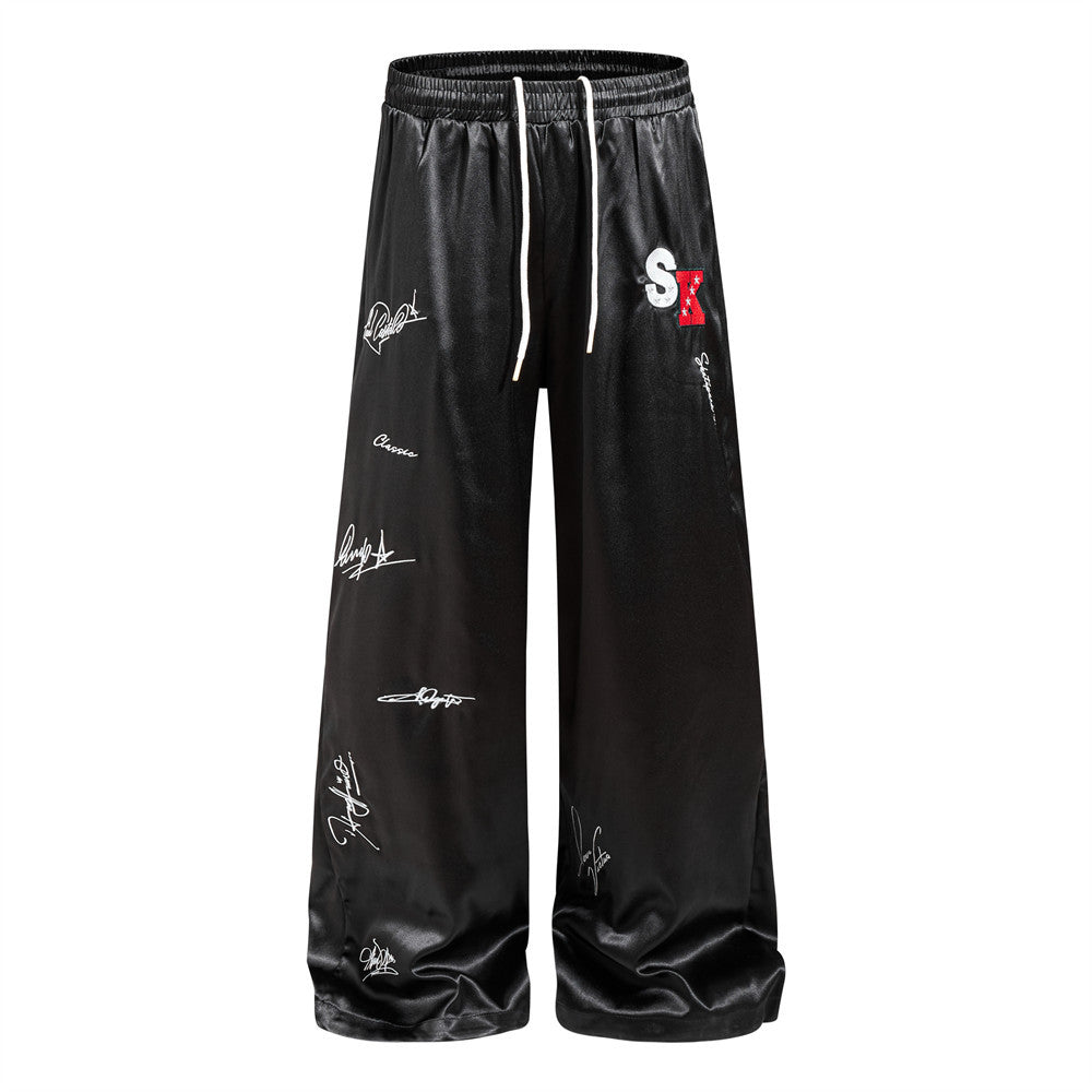 Men's wide leg black satin pants with all-over white signature script print and embroidered SK logo patch