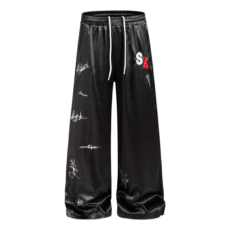 Men's wide leg black satin pants with all-over white signature script print and embroidered SK logo patch