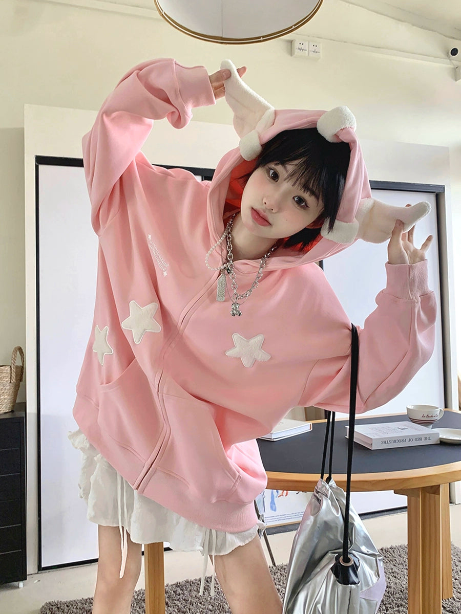 A person playfully poses indoors wearing the Seakoff Kawaii Star Elf Hoodie, featuring cute 3D ear details and soft star appliques. In the minimalistic, neutral-toned room, they stand near a table and shelf holding a metallic bag.