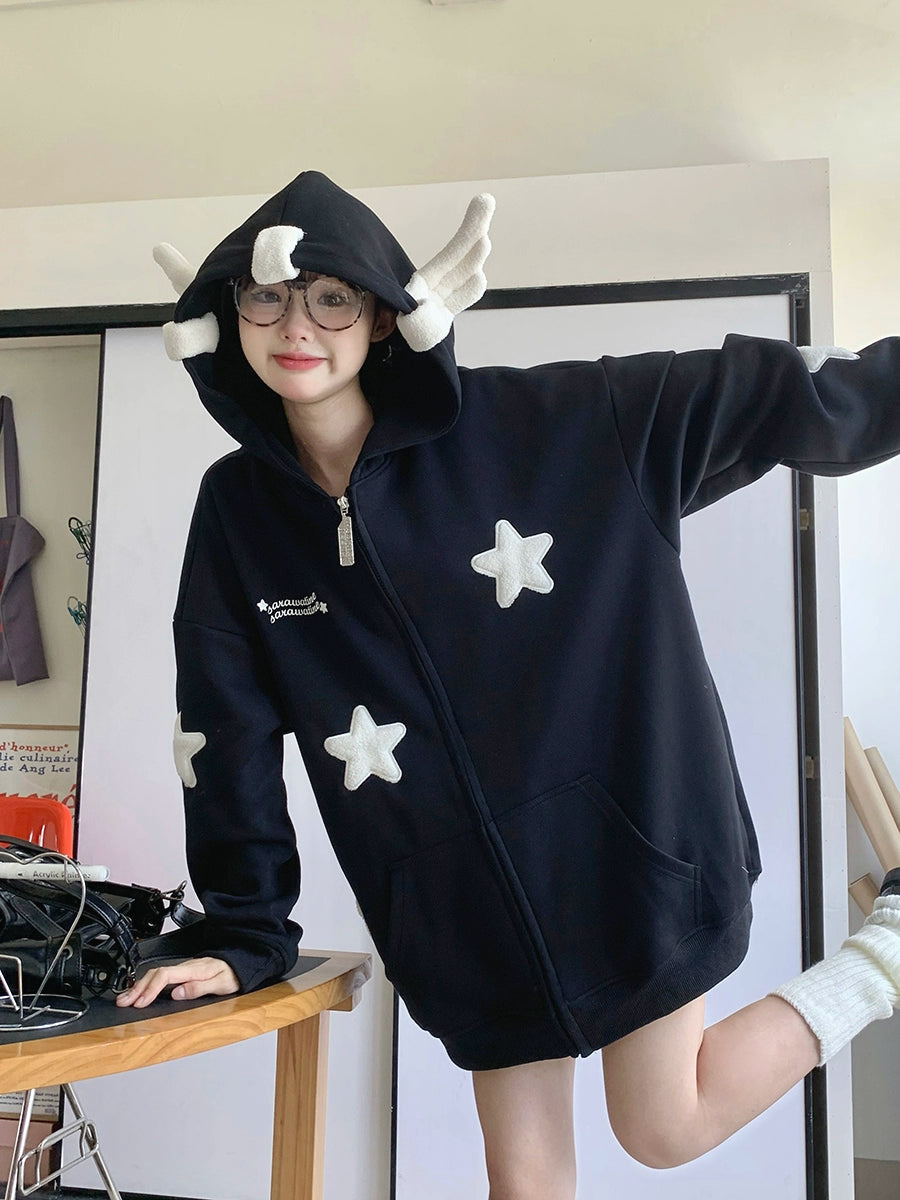 Indoors, someone models the Seakoff Kawaii Star Elf Hoodie—a whimsical piece with soft star appliques and 3D ear details. They smile, one leg slightly raised and one arm extended, as a table with various items lingers behind them.