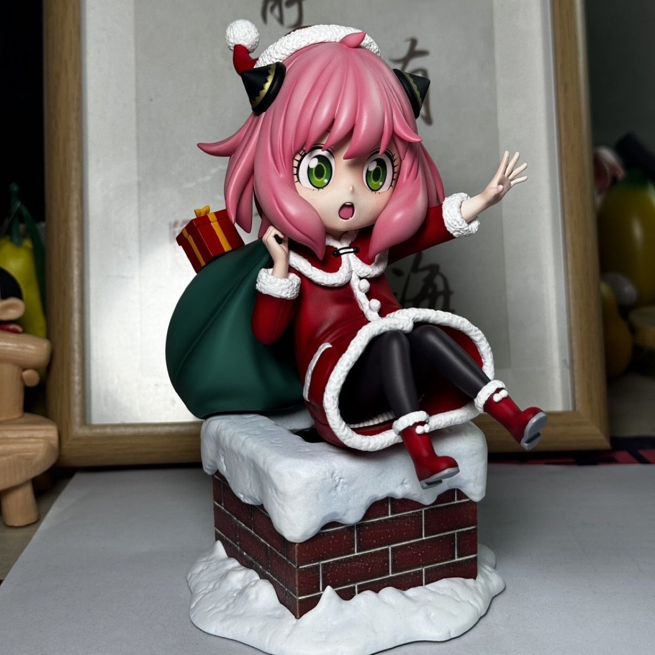 The Anya Forger Christmas Outfit Figure from Spy × Family features pink-haired, green-eyed Anya in a festive red and white attire. Seated on a snowy chimney with a green sack of gifts, it&