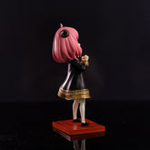 The Spy × Family Anya Forger PVC Figure features Anya Forger in a side view on a red base with pink hair, starry eyes, a black dress with gold accents, and white tights against a dark background. The figure measures 17 cm in height.