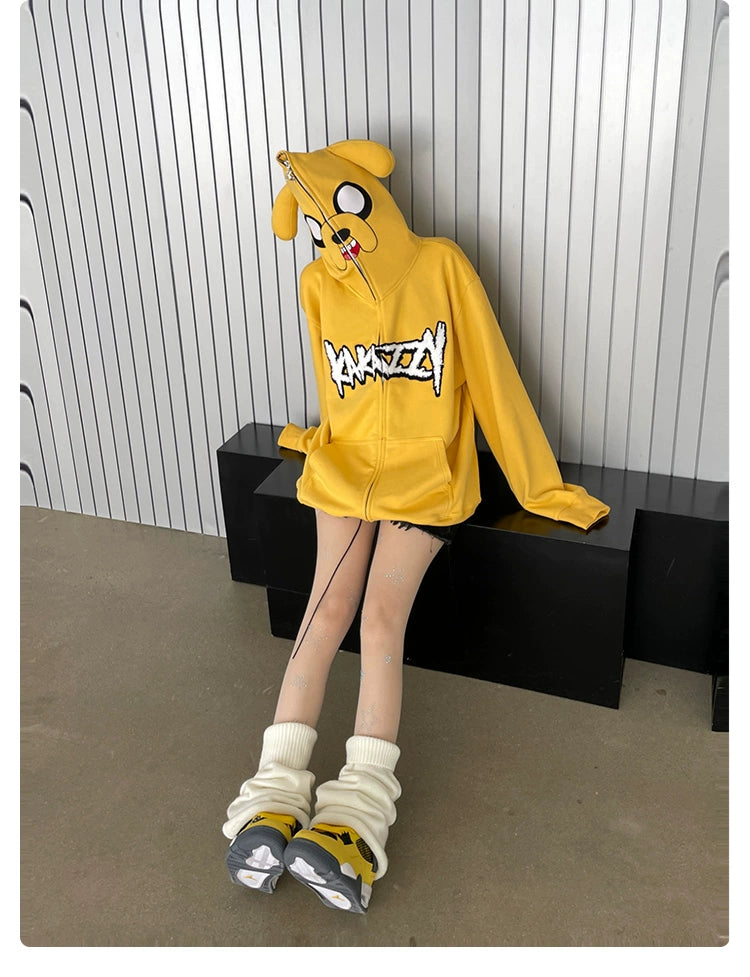 Someone in a Seakoff Kawaii Yellow Dog Hoodie, featuring ears and a cartoon face, sits on a black bench against a corrugated metal wall. The hoodie obscures their face as they wear white leg warmers and sneakers, with graphic designs on their thighs, capturing the anime streetwear style.