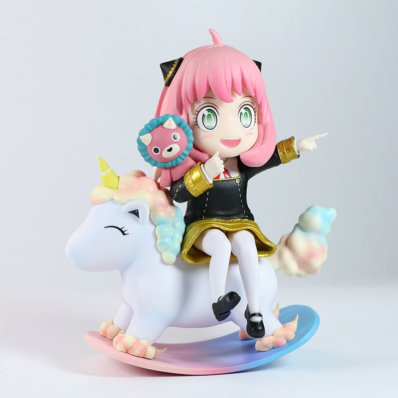 A Spy x Family Anya Forger PVC figure featuring chibi-style Anya with pink hair in a black and gold outfit on a pastel rocking unicorn. She holds a cat-faced object, has an excited expression, and points forward. The 15 cm figure includes an interchangeable expression.