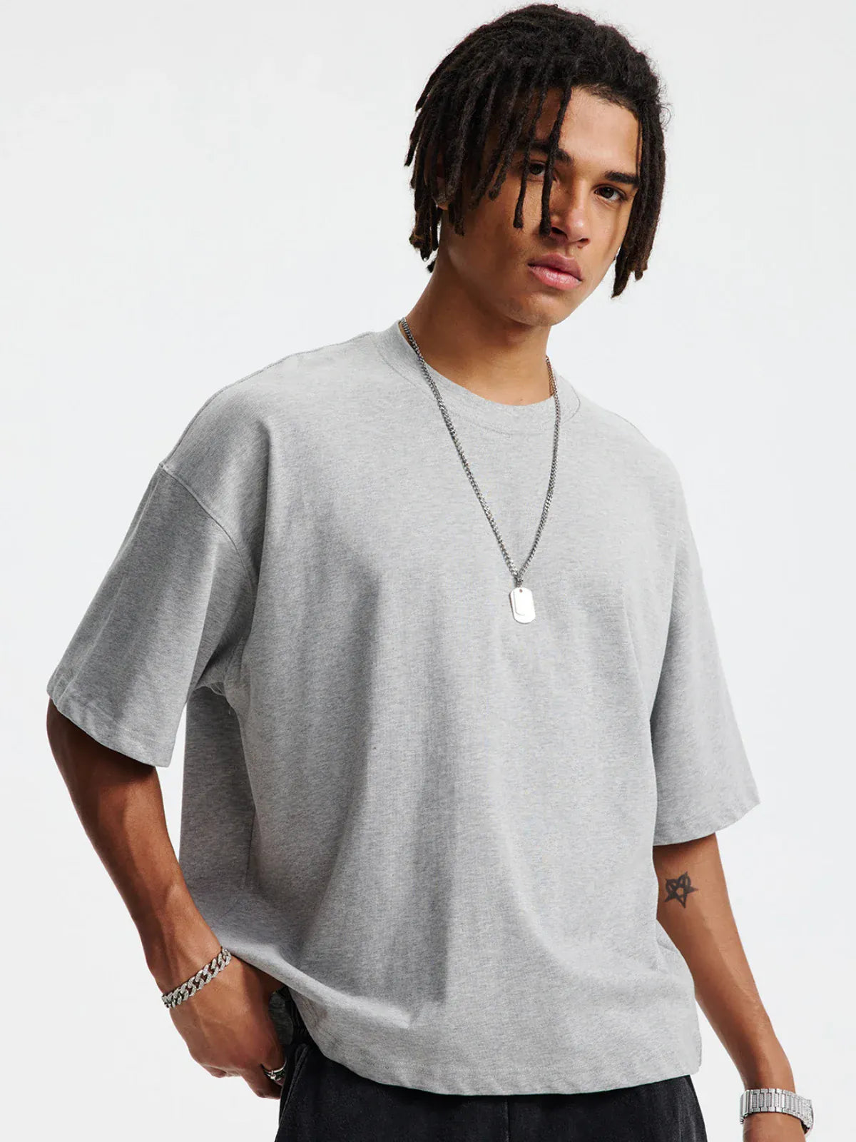 A person in a Custom Anime Character Heather Gray T-Shirt - Personalized 230GSM 100% Cotton Tee, black pants, a chain bracelet, watch, and rectangular pendant necklace stands against a plain background.