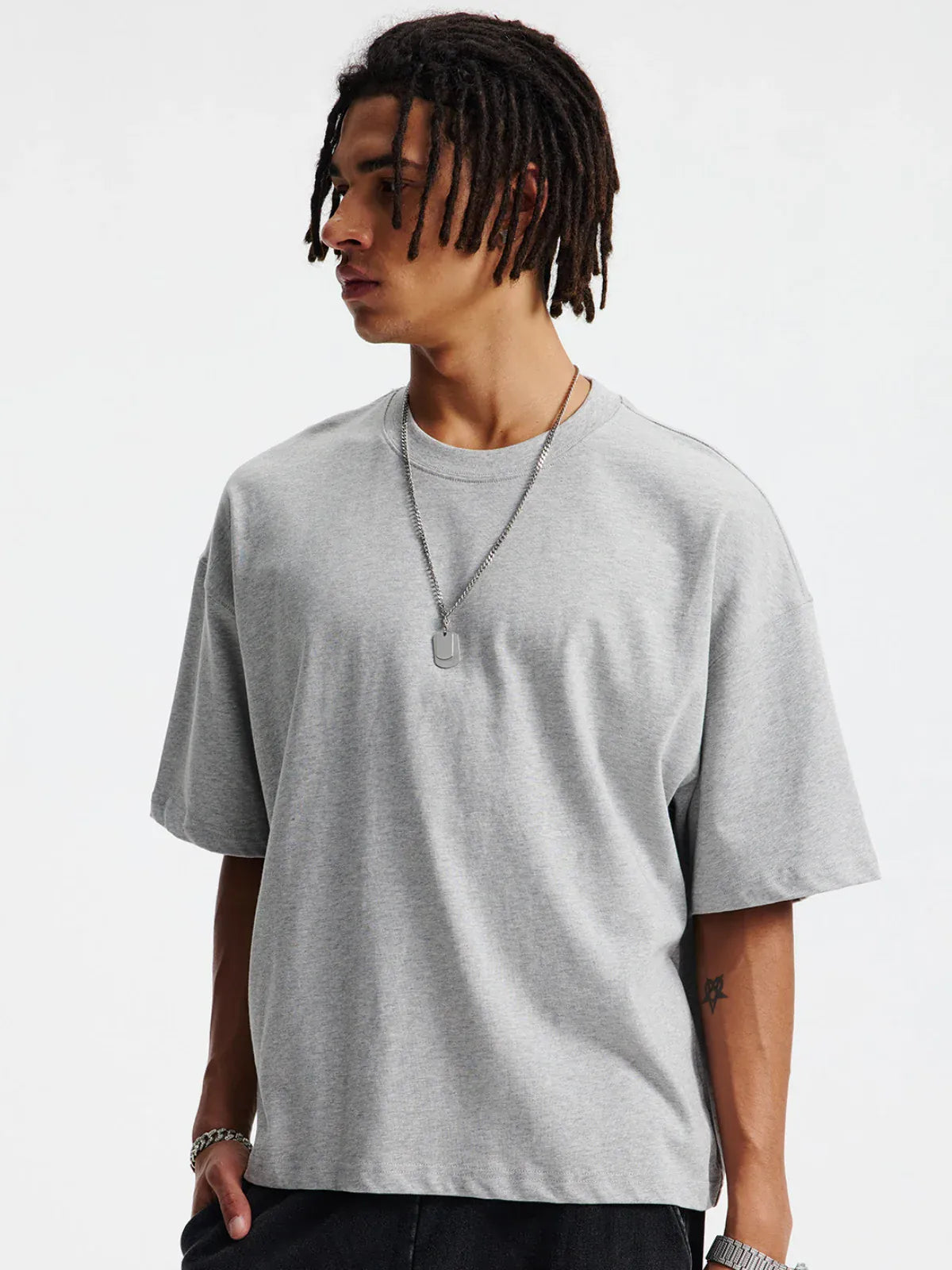 A person with medium-length dreadlocks wears a Custom Anime Character Heather Gray T-Shirt (230GSM 100% cotton), black pants, and a silver necklace while standing against a plain white background.