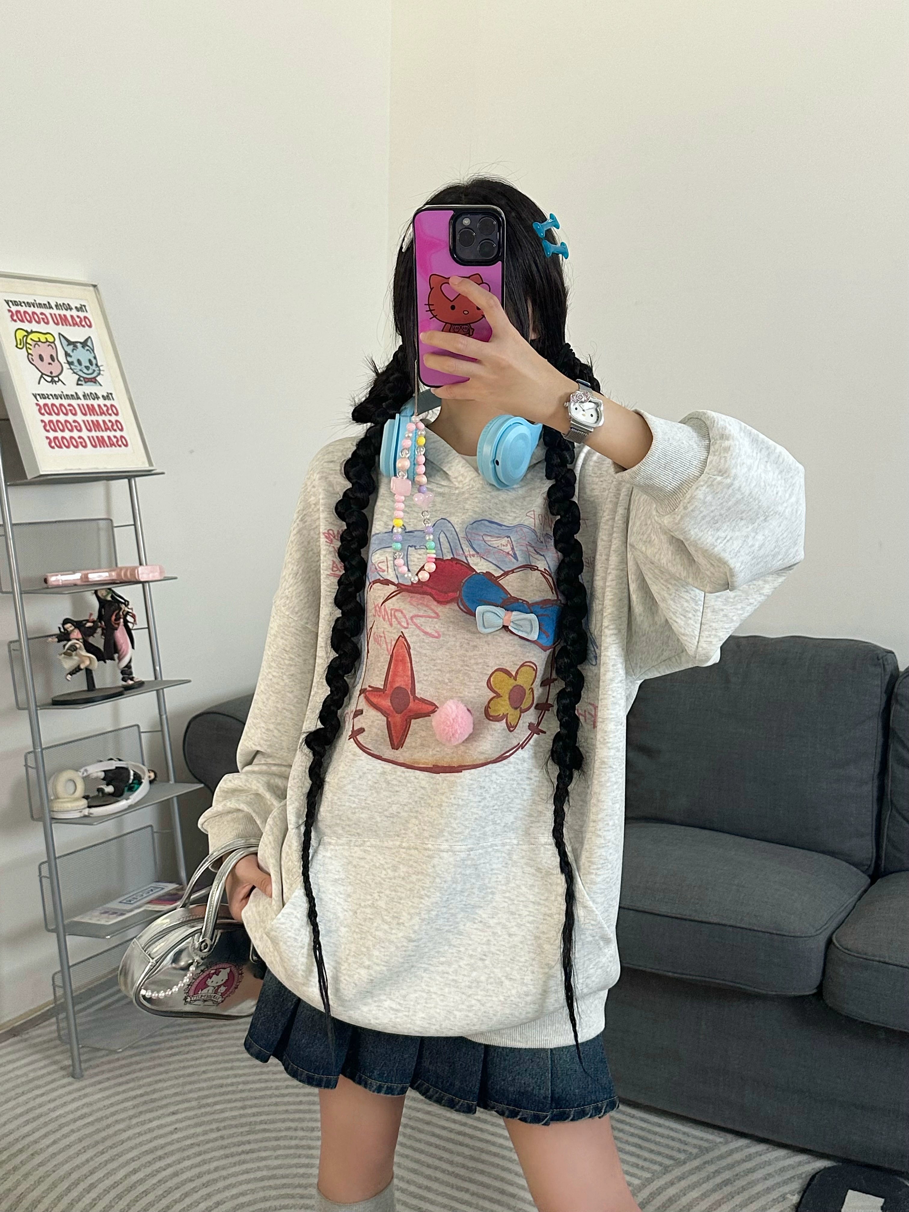 A person takes a mirror selfie in a Seakoff Kawaii Pom-Pom Crown Hoodie, paired with a denim skirt and long braided hair. Embracing the streetwear vibe, they hold a pink phone with a beaded charm while standing in a room featuring a gray couch and decorative items on shelves.