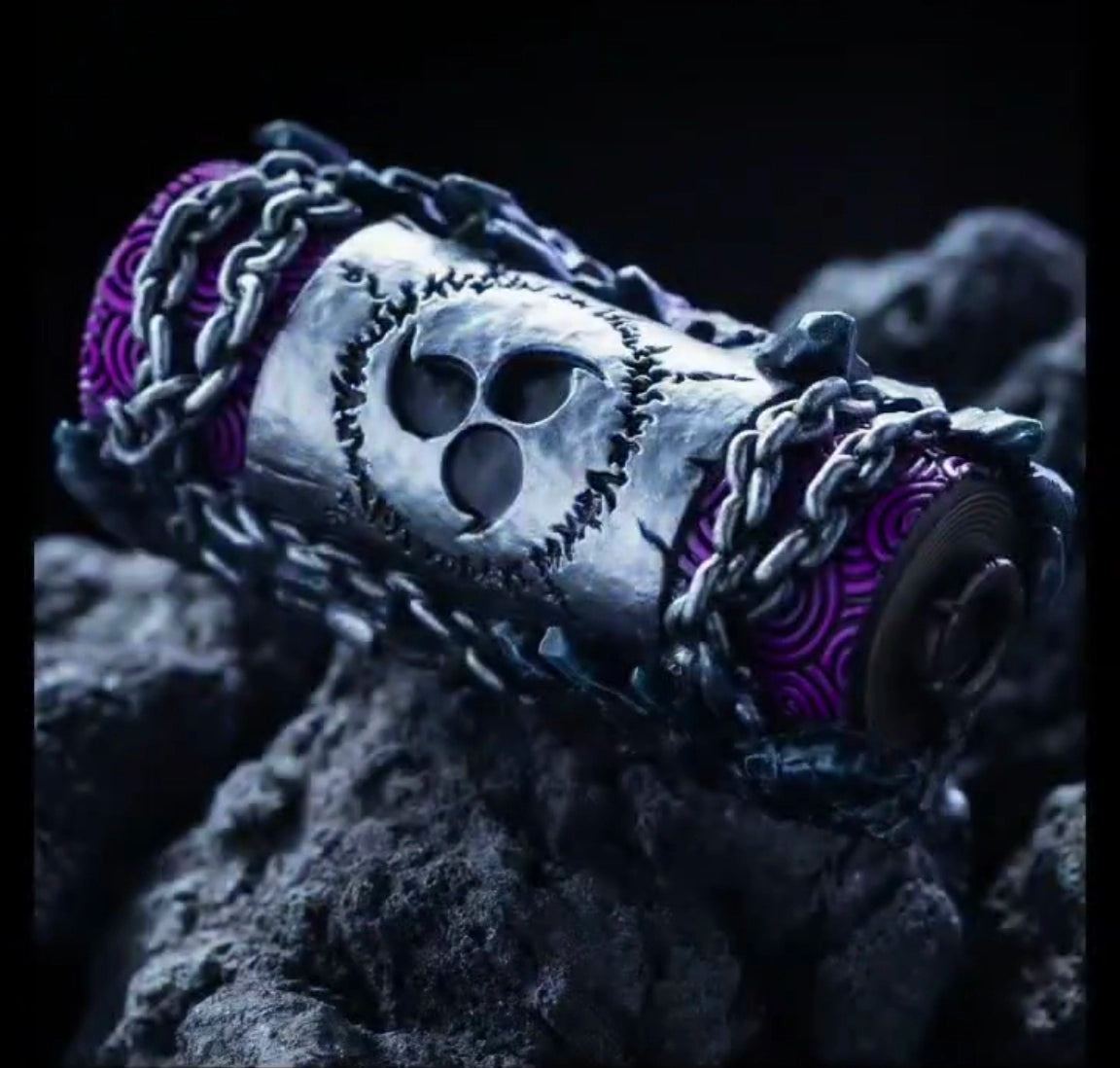 A cylindrical object, adorned with intricate chains and metallic designs, is placed on a rocky surface. It resembles the Sasuke Keycap Set – Curse Mark, Amaterasu, and Susanoo by Naruto with its purple patterns and central triskelion emblem.