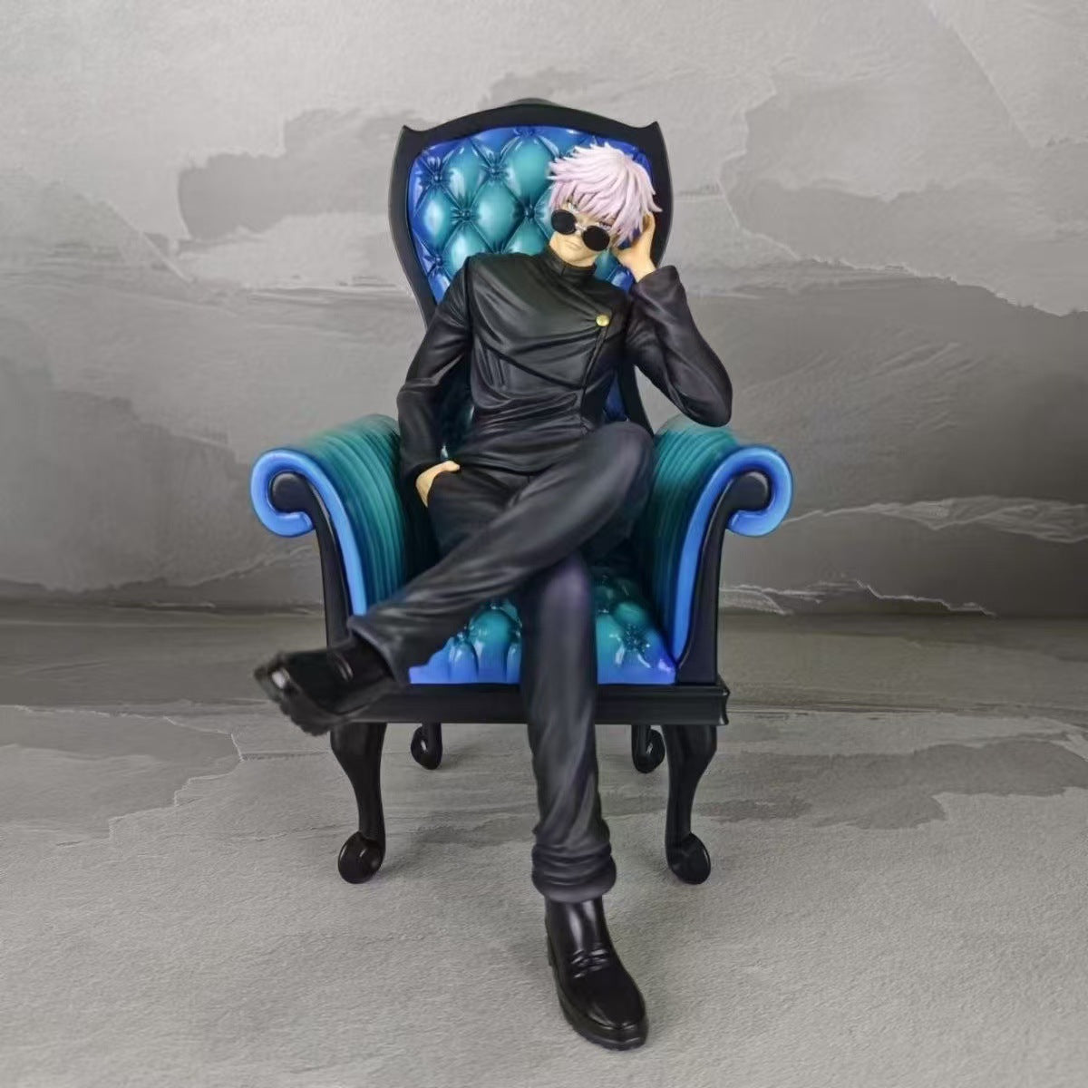 A Satoru Gojo figure from the Jujutsu Kaisen Satoru Gojo &amp; Suguru Geto Sitting Posture PVC Figure Set sits elegantly on a blue and black ornate chair, dressed in dark attire with light hair and sunglasses, one leg crossed against a gray background.