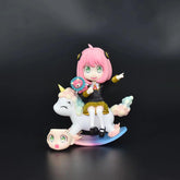 The 15 cm PVC figure of Anya Forger from Spy × Family features her with pink hair on a colorful unicorn rocking toy, pointing with one hand and holding a plush doll in the other, all set against a dark background. The figure also includes an interchangeable face expression.