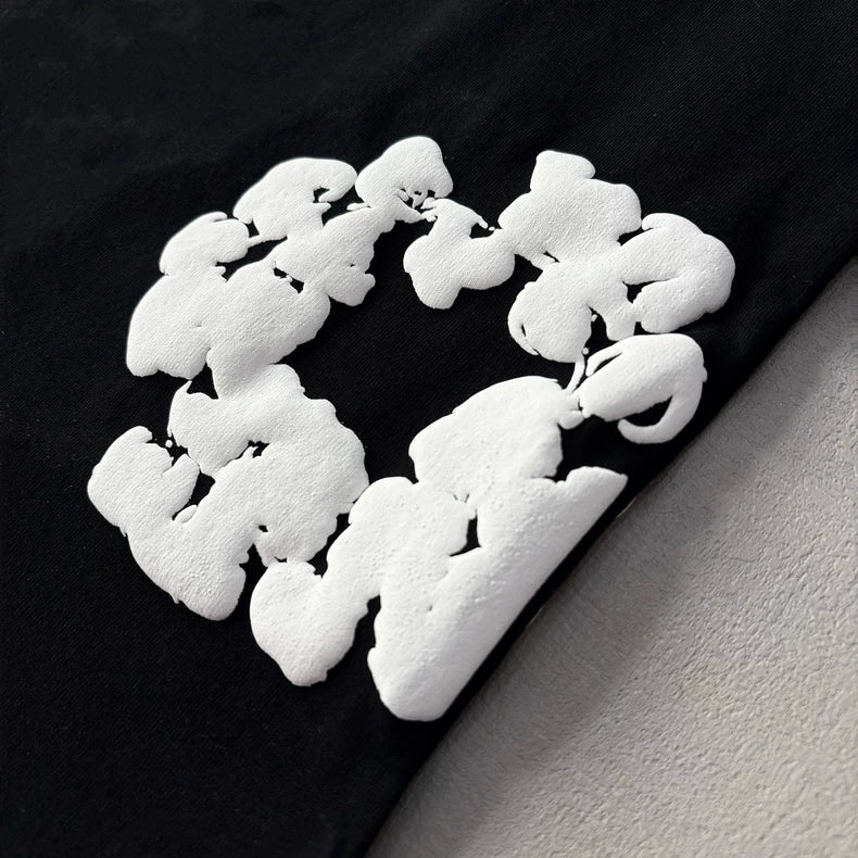 The Denim Tears Black Cotton Wreath All-Over Print T-Shirt features bold, white textured lettering in a circular design on black cotton. Raised, abstract 3D text gives this oversized streetwear tee a striking look against light backgrounds.