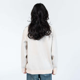 A person with long, wavy dark hair stands with their back to the camera, wearing the Dandadan Premium Turbo-Granny Possessed Knit Sweater in a unique jacquard design. They effortlessly pair it with blue jeans against a plain white wall, merging casual style with eclectic flair.