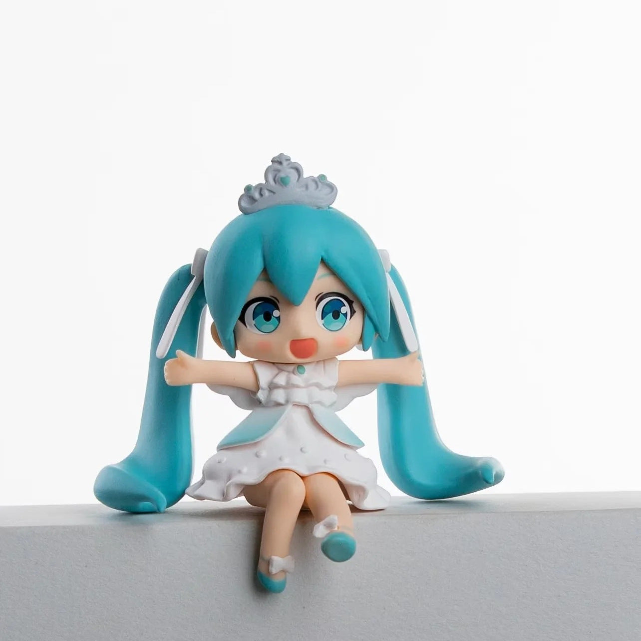 A 7.5cm PVC figurine from the Hatsune Miku Prayer Hugging Angel Princess set, featuring blue twin ponytails, a crowned angel princess in a white and blue dress with outstretched arms, sits on a ledge. The plain white background beautifully highlights its details.