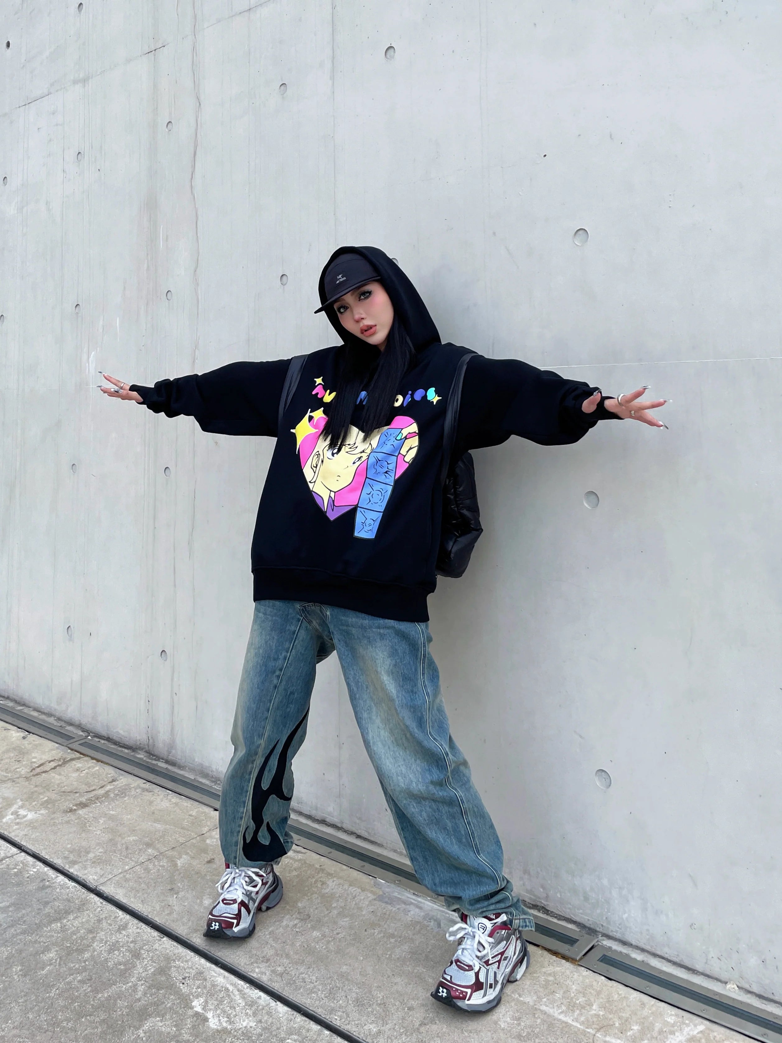 A person poses against a concrete wall wearing Seakoff's Retro Anime Hoodie - Human Voices Graphic Pullover with Drop Shoulder Design, baggy jeans, and sneakers. Sporting a black cap and nostalgic vibes, they extend their arms for that perfect retro flair.