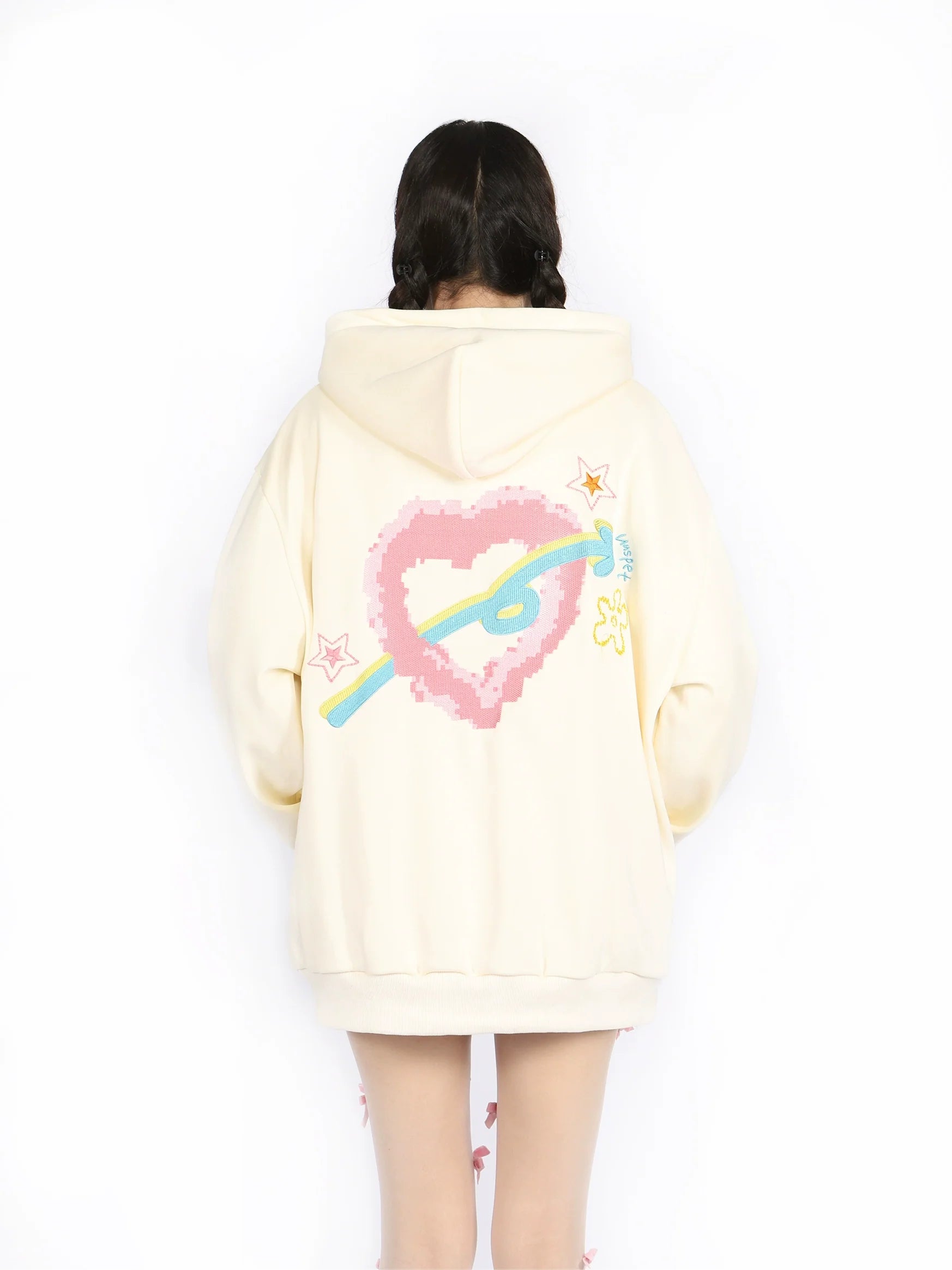A person with braided hair is wearing Seakoff's Kawaii Pastel Cartoon Hoodie, a cream-colored full-zip hoodie showcasing playful embroidered designs on the back, including a pink pixelated heart, colorful arrow, and small stars. Embracing kawaii fashion, the person faces away from the camera.