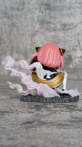 A pink-haired figure in gold and black attire, resembling the Spy x Family Anya Forger PVC Figure—9.5 cm Luffy Cosplay, Double Head Replacement, sits on a gray base. Pink and white flame accents surround the character, with cat-like ears facing away against a textured gray backdrop.