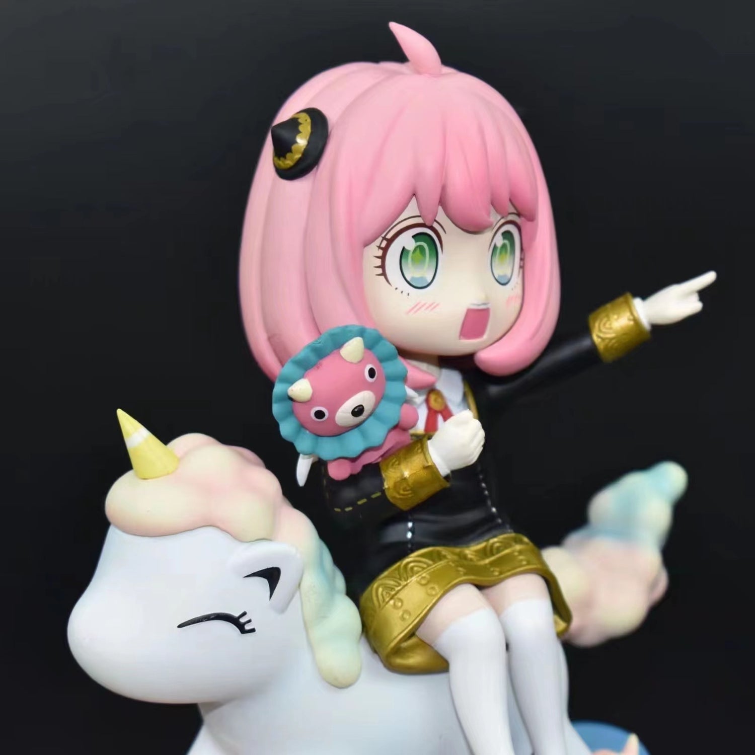 The Spy x Family Anya Forger PVC Figure showcases Anya with pink hair, perched on a unicorn with a vibrant mane and yellow horn. She has large green eyes, wears a black outfit with gold accents, and holds a plush toy. The unicorn looks content. Includes interchangeable face expression.