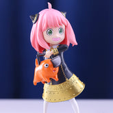 The Anya Forger Chainsaw Cosplay Figure, an 18CM PVC action collectible from Spy x Family, showcases pink hair, cat ears, and a black outfit with gold accents while holding an orange toy with large eyes and a wide grin in front of dark blue curtains.