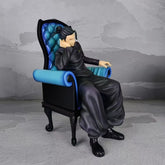 A figure from the Jujutsu Kaisen Satoru Gojo & Suguru Geto Sitting Posture PVC Figure Set features Suguru Geto in a dark outfit on a blue-upholstered high-backed chair, with their head resting on one hand against a textured gray background.