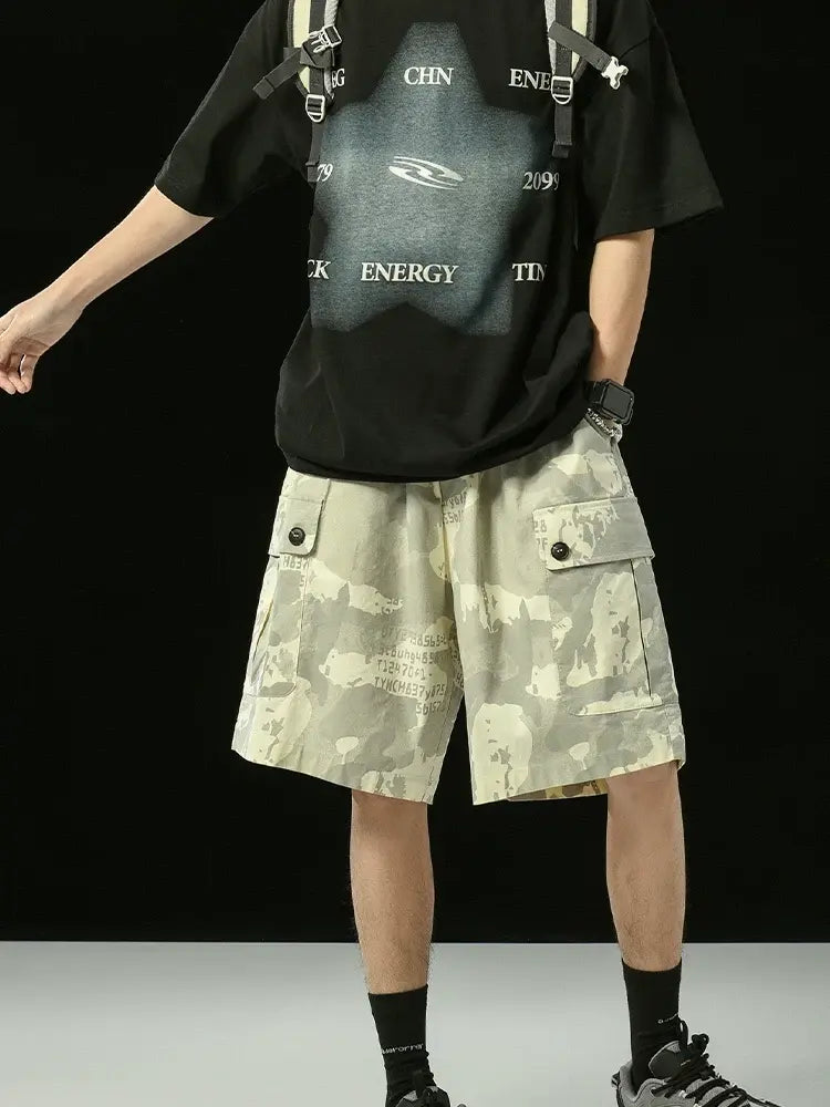 A person in a black graphic t-shirt, black socks, sneakers, and Men's Grey Camo Cargo Shorts with Button Flap Pockets and Elastic Waist stands against a dark background. Their head is out of frame, carrying a light-colored backpack.