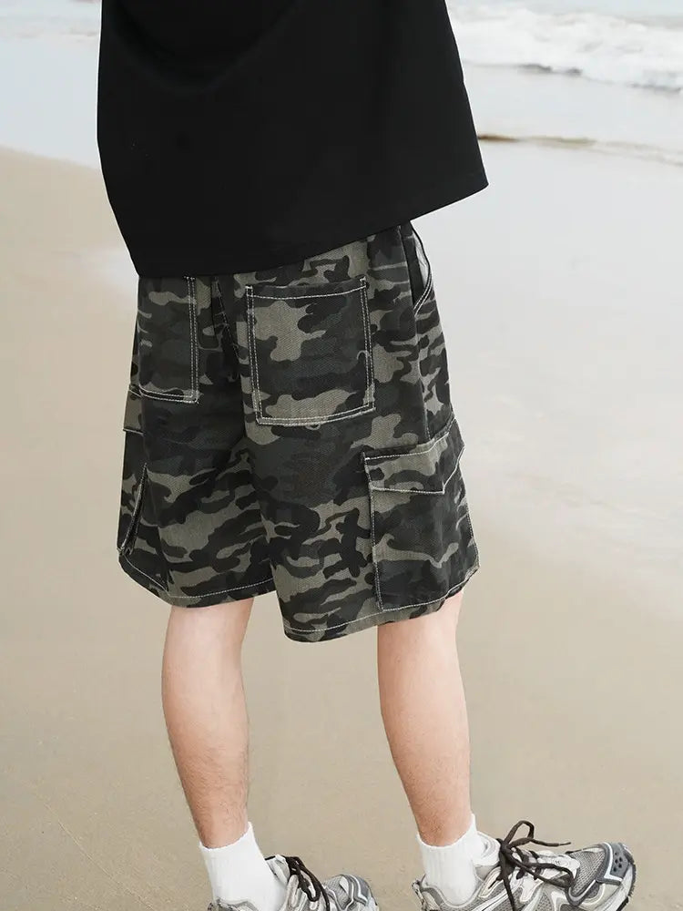 A person stands on the beach wearing Men's Baggy Camo Cargo Shorts with Elastic Drawstring Waist and Flap Pockets, a black shirt, white socks, and gray sneakers. Only their legs and part of their torso are visible, ocean behind them.