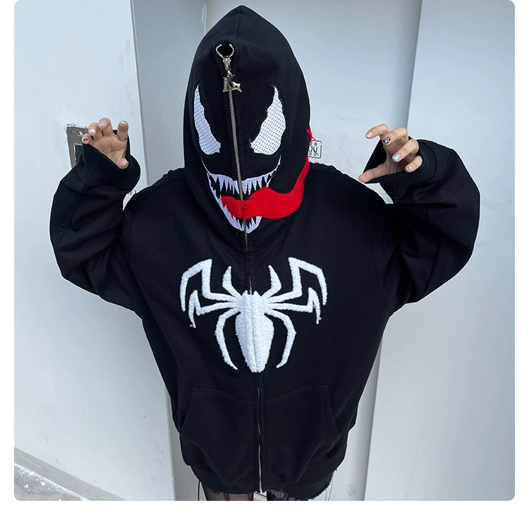 A Marvel fan wears a Seakoff Venom-Inspired Hoodie, crafted from premium 400GSM cotton. The black hoodie features a signature tongue with eyes, teeth, and a spider logo on the hood, embodying the popular comic character against a plain, light-colored wall.