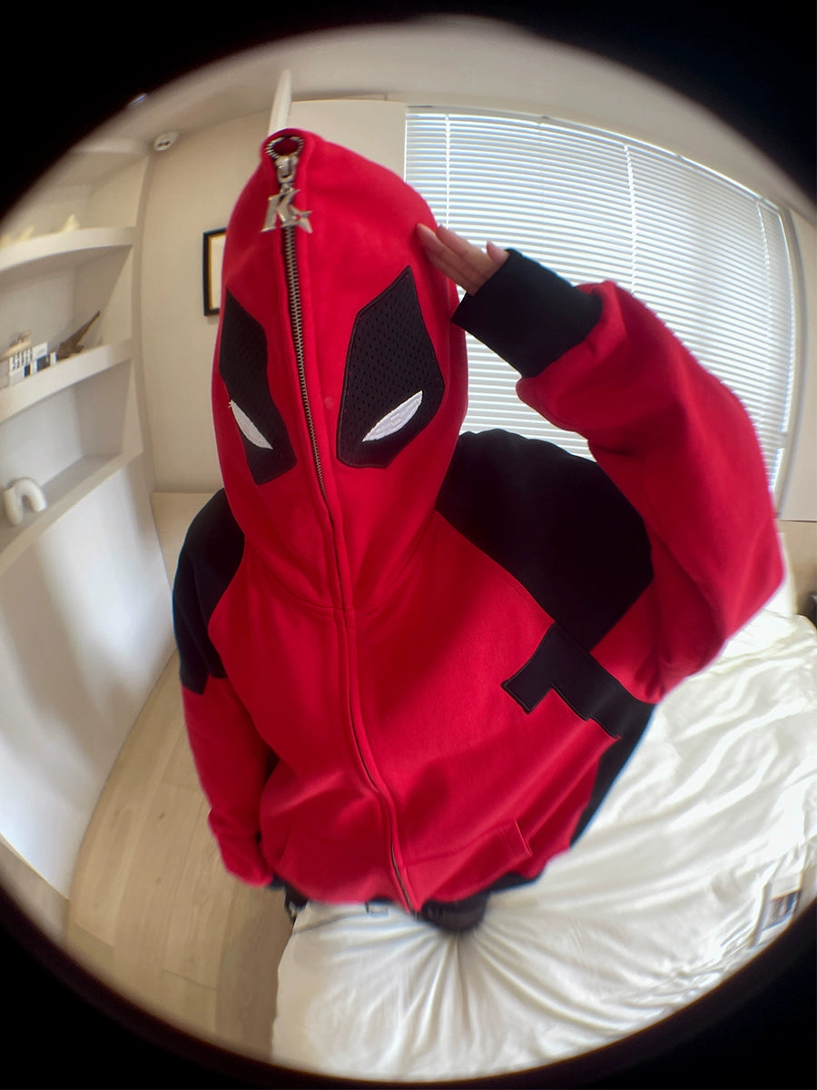 A person in a Seakoff Deadpool-Inspired Hoodie, featuring an oversized red and black design that resembles a superhero mask, sits indoors. The hood obscures their face as they salute with their right hand, while the fisheye lens effect adds an edgy touch to the dynamic fit.