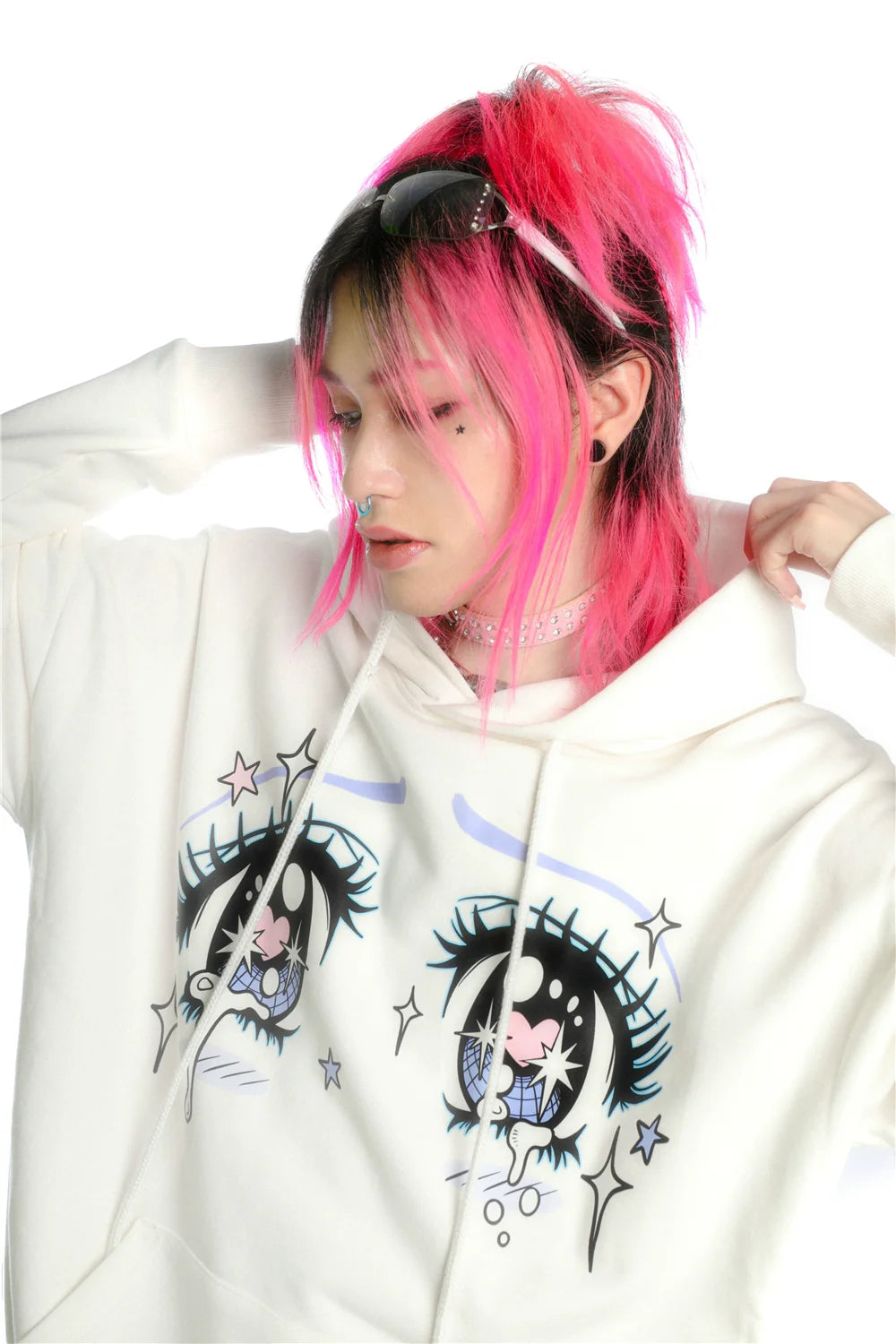 With bright pink hair and embodying the streetwear vibe, a person wears Seakoff's Kawaii Aesthetic Crying Eyes Hoodie—a white anime-style pullover with sleeve graphics—and sports sunglasses on their head, a pink choker, and a nose piercing while looking down with one hand behind their head.