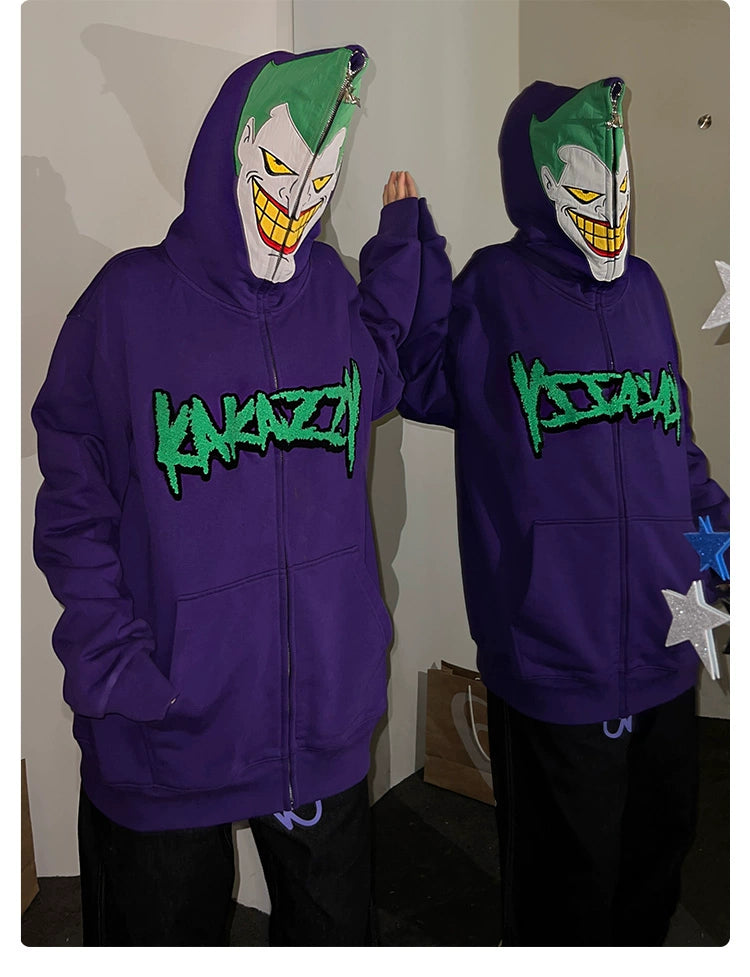 Two people wearing Seakoff Joker-Inspired Hoodies, 400GSM oversized purple zip-up sweatshirts with green text and face masks featuring a menacing grin and green hair. They stand side by side, each with one hand raised, alongside a paper bag and decorative stars—ideal for supervillain fans.