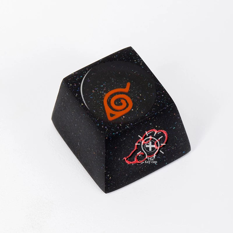 The Naruto Elements Keycap Set by Naruto features a black resin keycap with an orange swirl design inspired by the Hidden Leaf Village emblem. The side displays a red outline of a hand holding a key on a speckled surface.