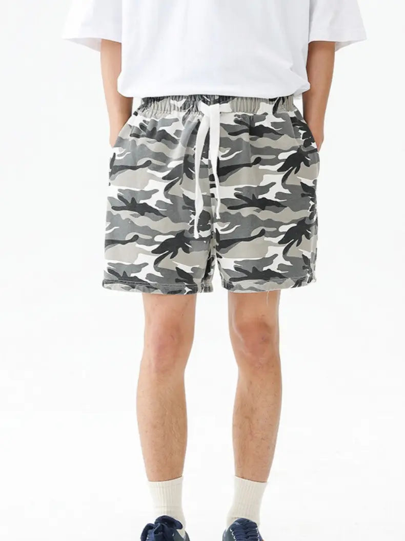 A person stands with hands in pockets against a plain white background, wearing a white t-shirt, navy sneakers, and Men's Urban Camo Cotton Drawstring Shorts - Gray Camouflage Relaxed Fit with a white drawstring.