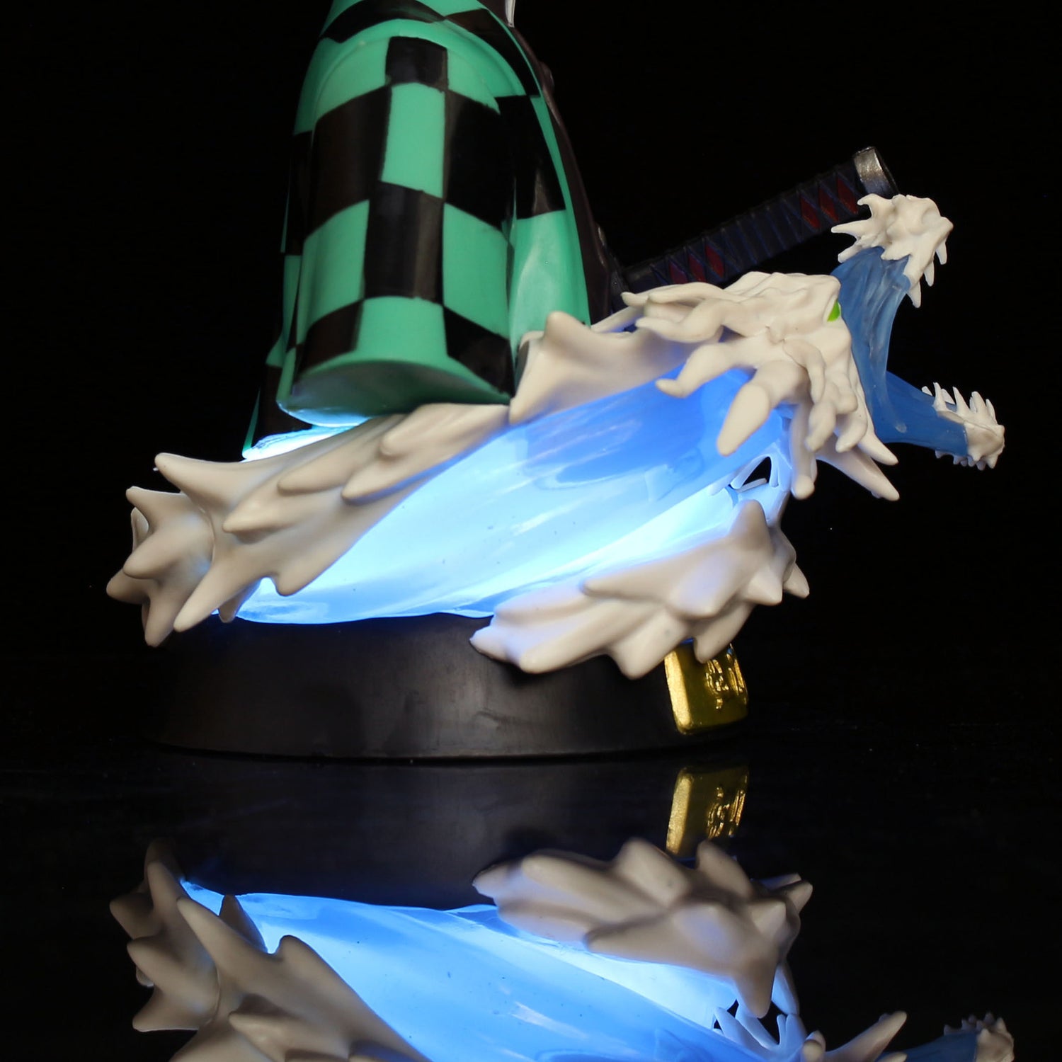 The Demon Slayer Tanjiro Kamado GK Half-Body Action Figure depicts Tanjiro in a green and black checkered cloak wielding a sword, surrounded by a blue dragon-like swirl with white accents, all reflected on a shiny black base. The 15cm collectible includes an LED light effect.