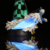 The Demon Slayer Tanjiro Kamado GK Half-Body Action Figure depicts Tanjiro in a green and black checkered cloak wielding a sword, surrounded by a blue dragon-like swirl with white accents, all reflected on a shiny black base. The 15cm collectible includes an LED light effect.