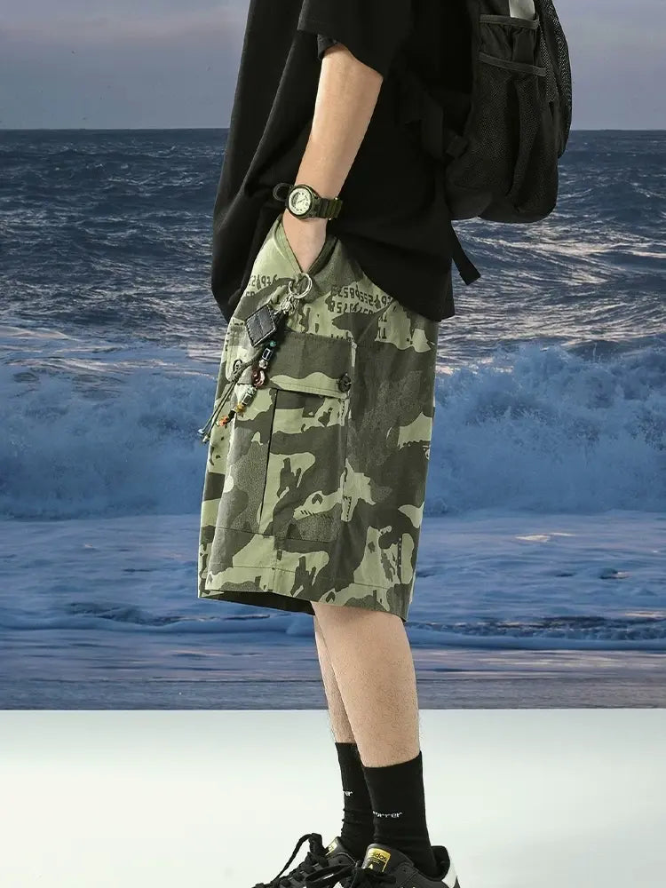 A person by the ocean wears a black t-shirt, Men's Green Camo Cargo Shorts with Drawstring Waist and Button Flap Pockets, black socks, black sneakers, a black backpack, and a watch for a military-inspired streetwear look.