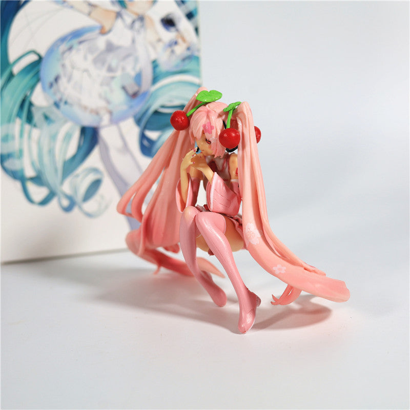 The Hatsune Miku Cherry Sitting Pose 11cm PVC Figure features the character with long pink hair and cherry accessories, gracefully cross-legged in a charming pink outfit with cherry blossoms, set against a vibrant illustration.