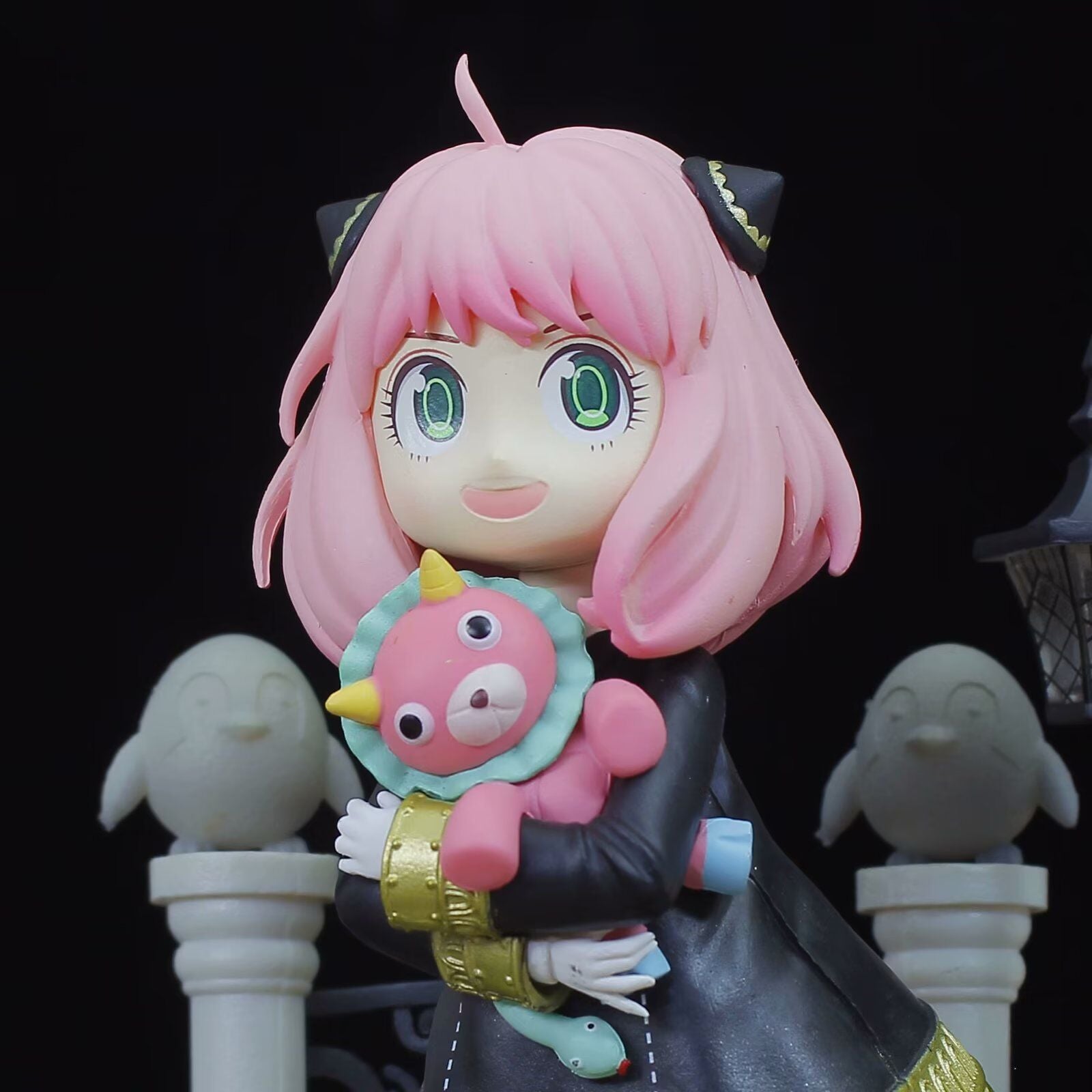 The Spy x Family Anya Forger 19 cm PVC Figure features a girl with pink hair and cat ear headbands holding a horned plush toy, large green eyes, and a dark coat with gold accents, standing before ghost-like figures and part of a fence—ideal for Spy x Family fans.