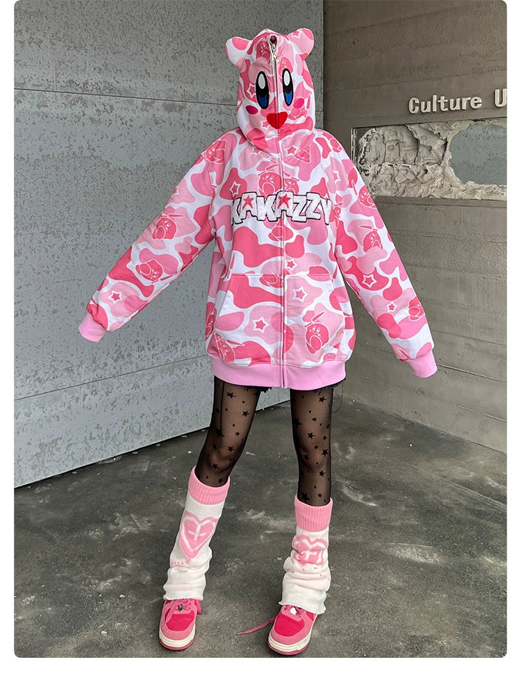 By a gray wall, a person wears the Seakoff Pink Kirby-Inspired Hoodie, featuring a camo design and 3D ears. Black star-patterned tights, white leg warmers, and pink sneakers complete their playful outfit.