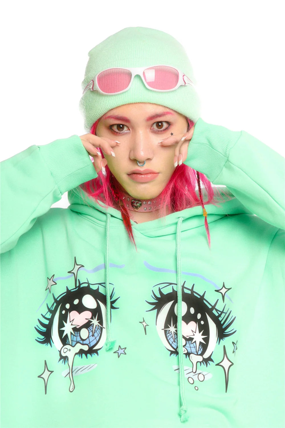 A person with pink hair strikes a pose, wearing Seakoff's Kawaii Aesthetic Crying Eyes Hoodie in light green. Their streetwear look is complete with a matching beanie, pink sunglasses, colorful makeup, and a choker necklace as they hold the hood with both hands.