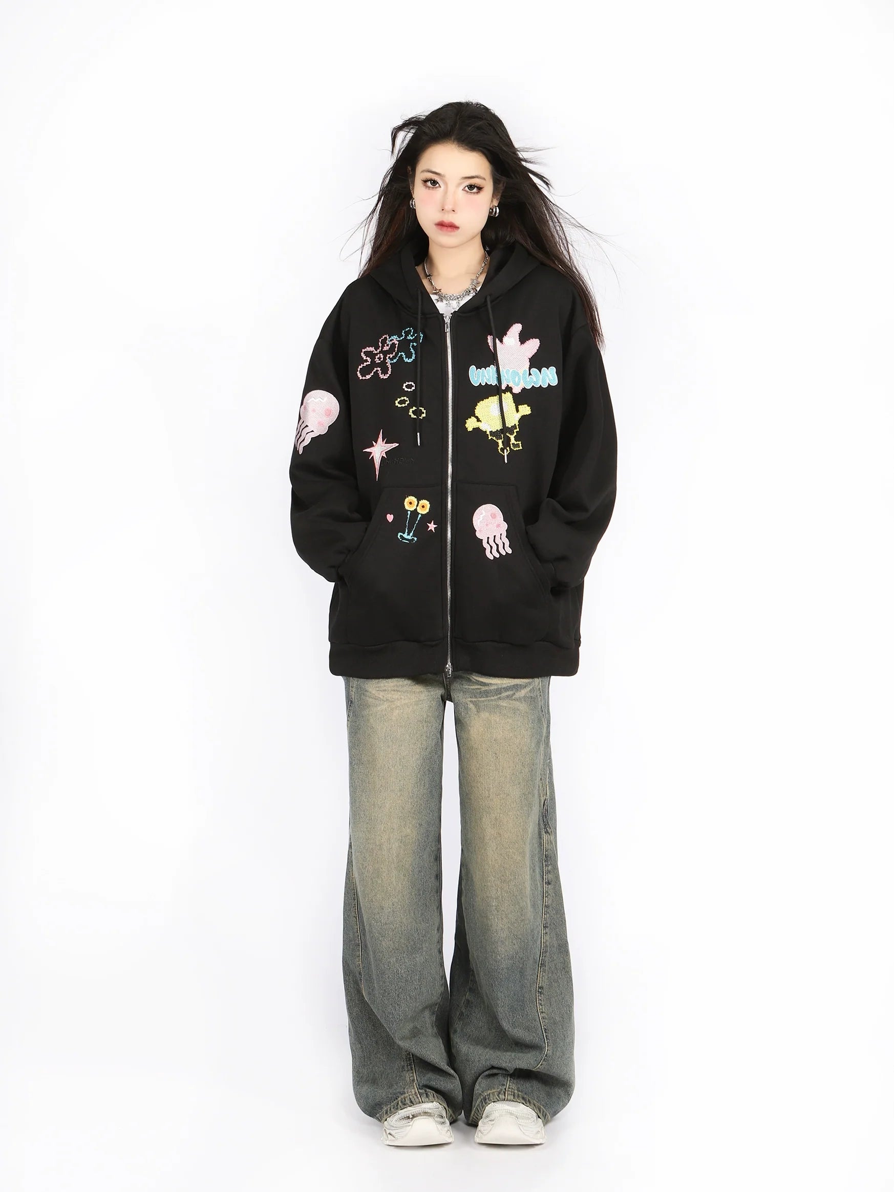 Against a plain white background, a person showcases their anime-inspired fashion with long dark hair, white sneakers, and baggy jeans while wearing Seakoff's Kawaii Pastel Cartoon Hoodie—a cute full-zip hoodie featuring various abstract shapes and playful embroidered designs.