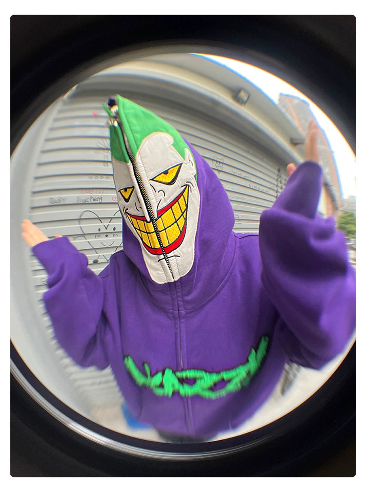 A person wearing a Seakoff Joker-Inspired Hoodie, featuring a purple and green design with a cartoonish smile, raises their hands outdoors against graffiti-covered metal shutters. Captured through a fisheye lens, it&