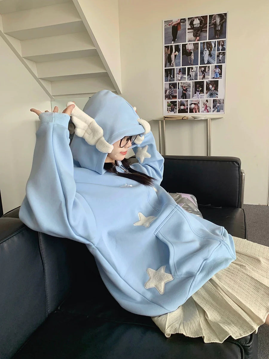 Lounging on a black couch, a person sports Seakoff's Kawaii Star Elf Hoodie, featuring an oversized fit with 3D ears and white star appliques on light blue fabric. As they adjust the hood, a photo-filled poster decorates the space behind them.
