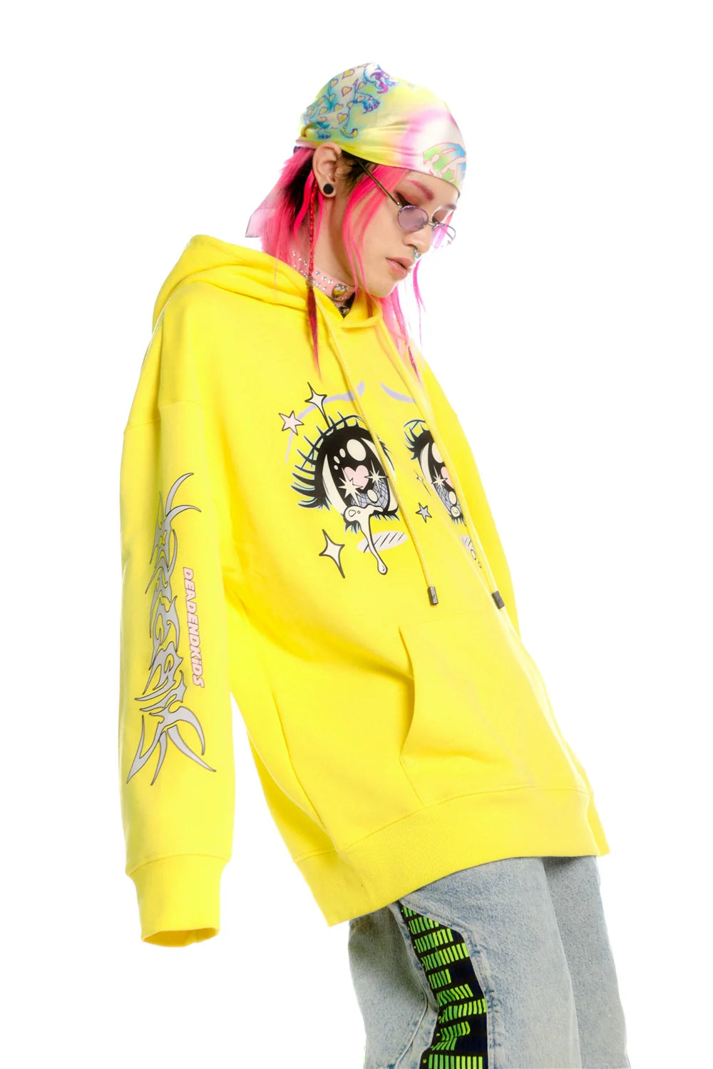 Wearing Seakoff's bright yellow Kawaii Aesthetic Crying Eyes Hoodie—a pastel and dark anime pullover with sleeve graphics—paired with light blue jeans featuring green patterns and a colorful bandana, this individual rocks bold streetwear. Their pink hair and sunglasses pop against the white background.