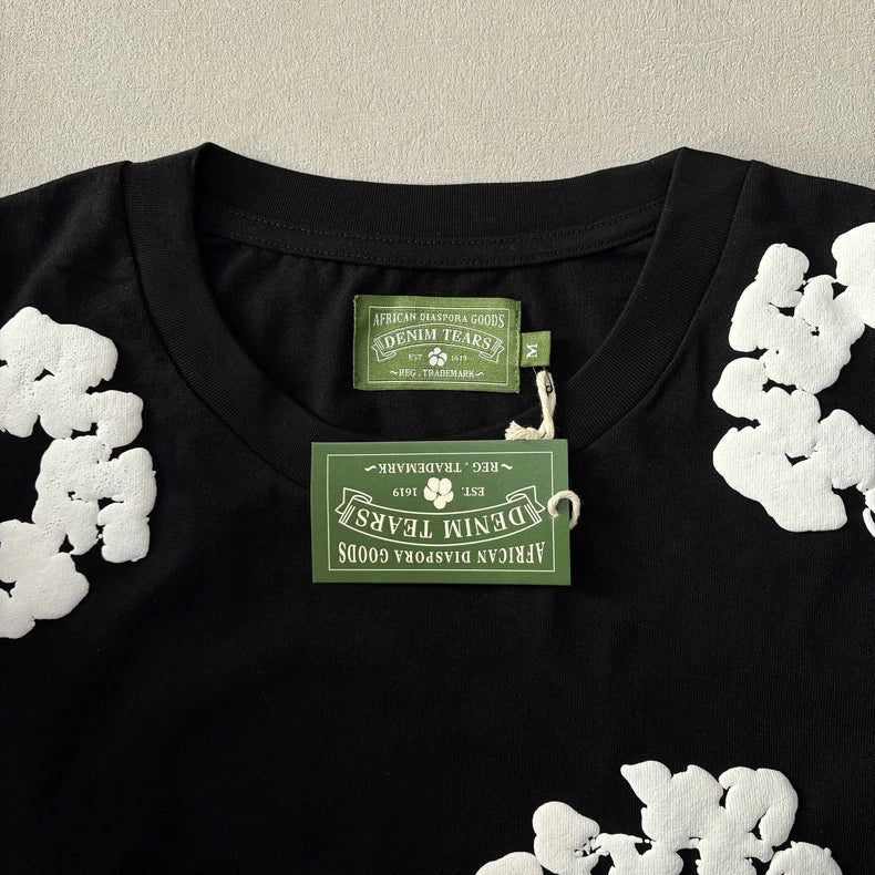 The Denim Tears Black Cotton Wreath All-Over Print T-Shirt features white floral graphics near the shoulders, two green “Denim Tears African Diaspora Goods” tags at the collar, and is displayed flat on a light surface.