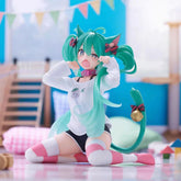 The Kneeling Cat-Ear Hatsune Miku 11cm PVC Figure from Hatsune Miku features a playful pose with long teal hair, a white shirt, and striped pink socks. Set on a shiny floor, soft-focus details like flags and cushions create a whimsical playroom backdrop.