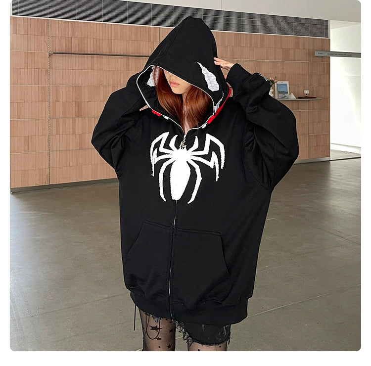 A person dons a black Seakoff Venom-inspired hoodie made from 400GSM cotton, featuring a white spider and signature tongue logo on the front. They stand in a spacious room with wooden walls, hood up, stylishly pairing it with patterned leggings—a must-have for Marvel fans.