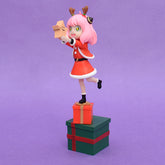 The 18cm Anya Forger Christmas PVC Figure from Spy × Family features a pink-haired character in a red Santa outfit with reindeer antlers, balancing on one leg on stacked gift boxes, holding an open box against a festive purple background.