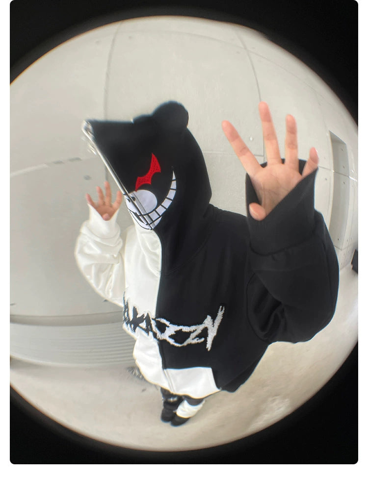 A person wearing a Seakoff Monokuma-Inspired Hoodie, featuring a smiling face and red eyes on the hood, poses with one hand up and the other on the hoodie. Against a plain background, this fisheye lens shot captivates Danganronpa fans.