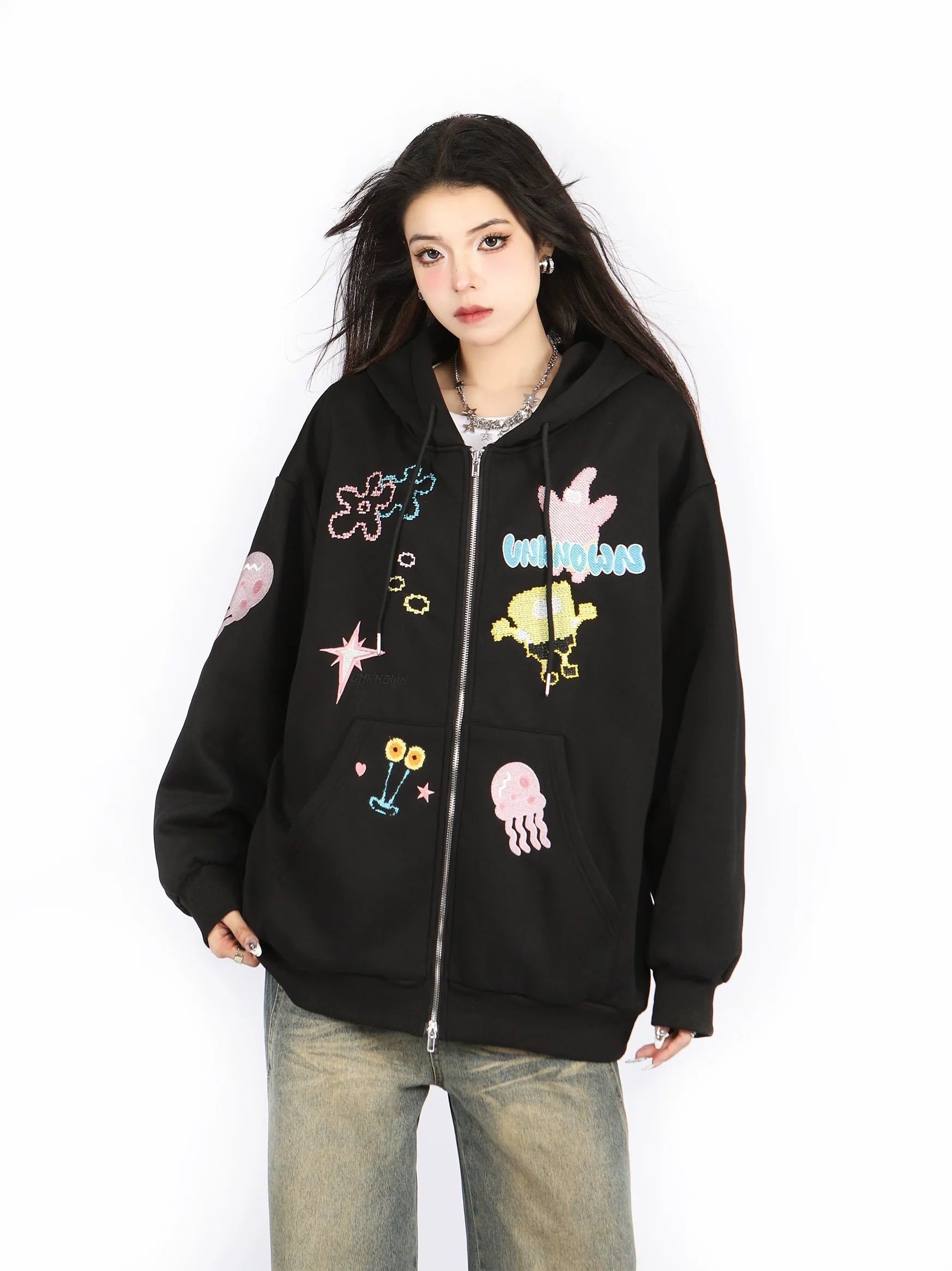 A person wears Seakoff's Kawaii Pastel Cartoon Hoodie, a cute black zip-up with playful embroidered stars and jellyfish. Their long dark hair complements loose-fitting blue jeans against a plain white background.